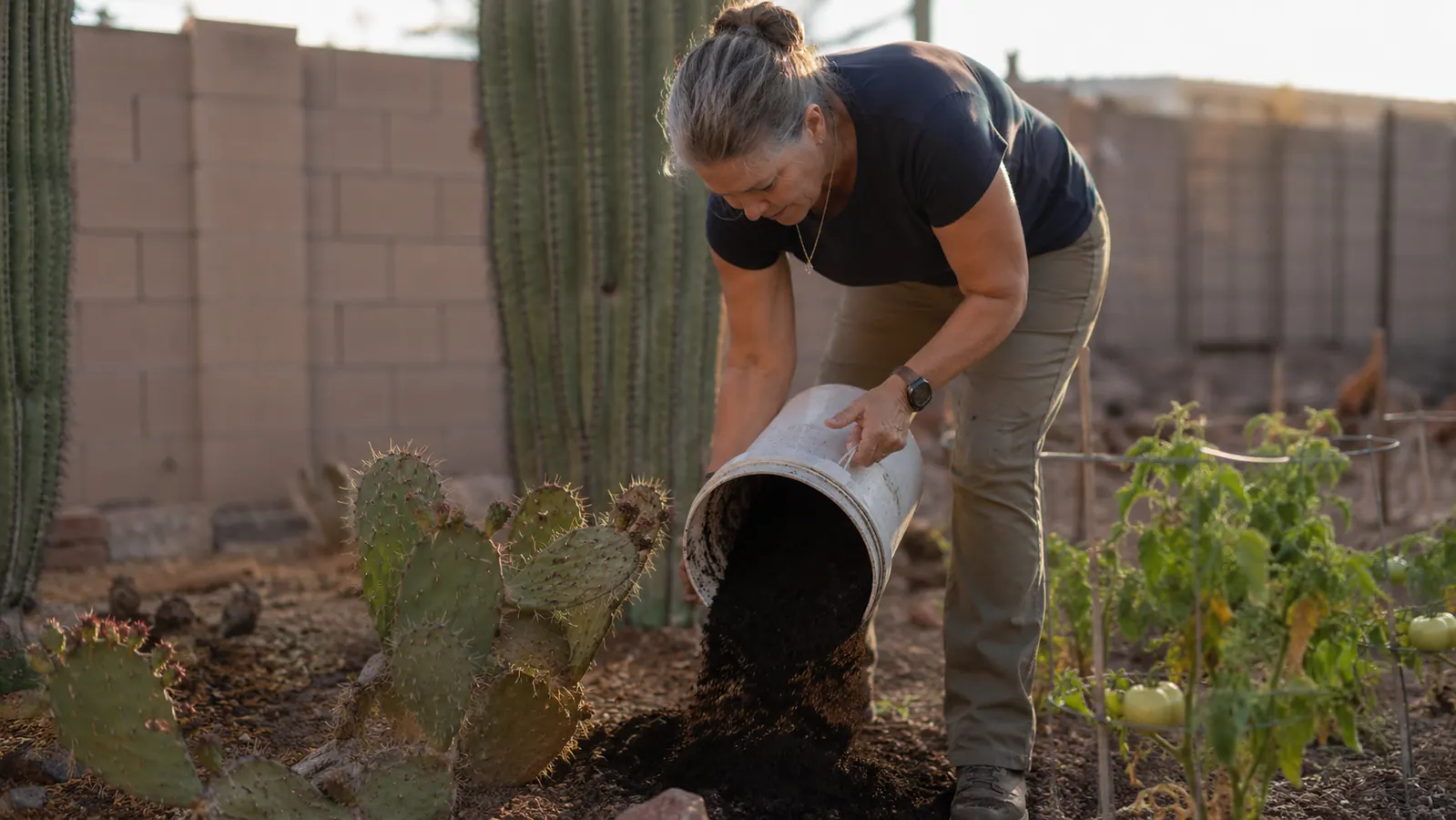 Woman in her fifties emptying dried compost grounds from a countertop composter bucket into a garden bed